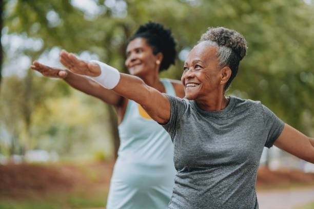 Two older women exercise outdoors, smiling while stretching their arms. The scene emphasizes joy and fitness in a natural setting, promoting healthy living.