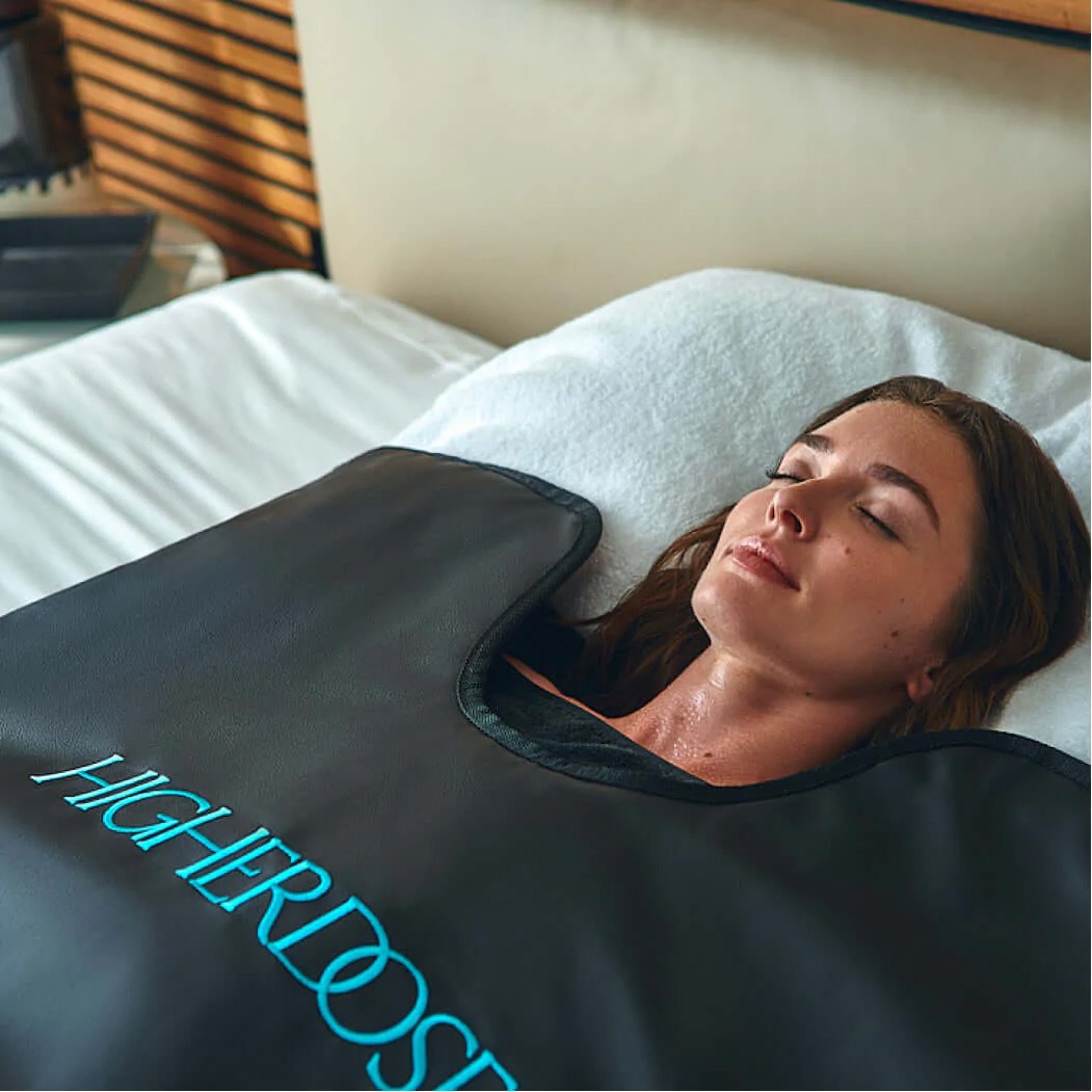 A woman relaxes on a bed wearing a black body wrap labeled "HigherDose," promoting wellness through heat therapy.