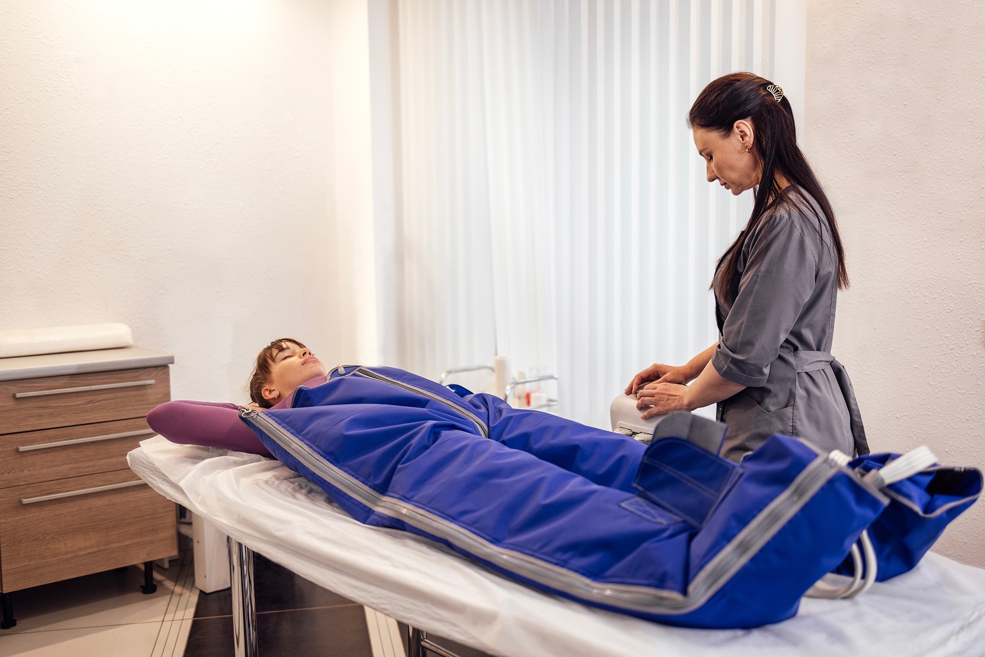 A woman in a blue body wrap lies on a treatment table while a therapist prepares a device for a wellness treatment in a spa setting.